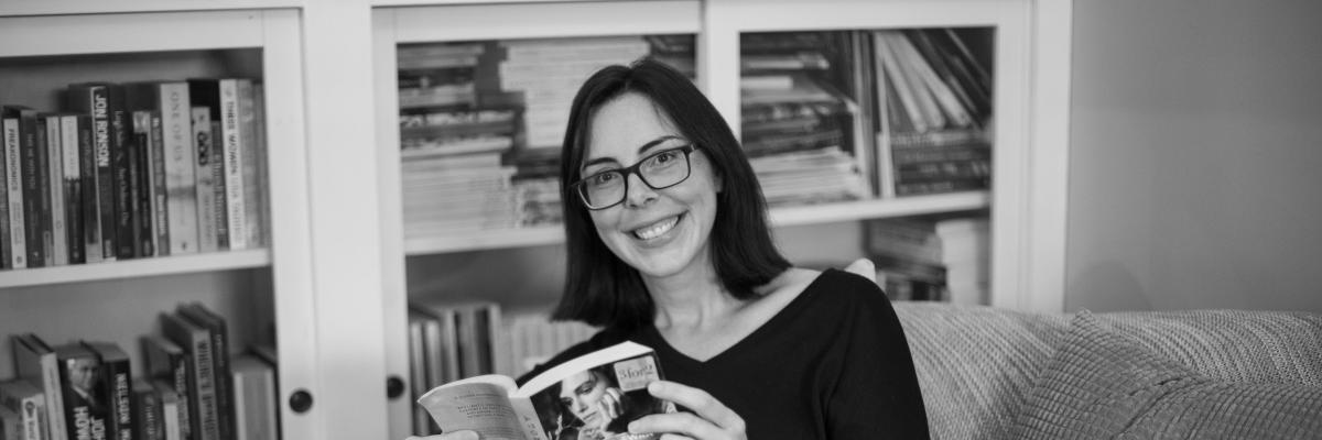 A black and white photo of a woman with a dark bob and glasses, sitting in a chair in front of a book shelf and reading a book.