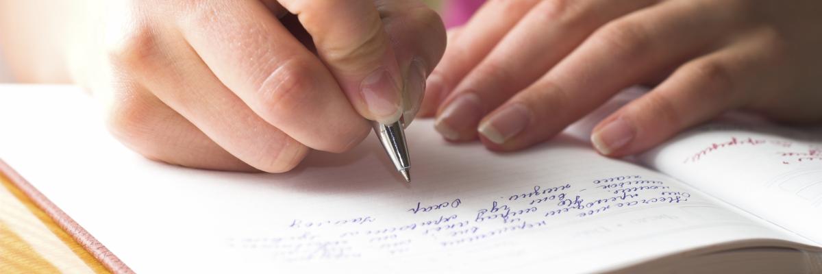 A person wearing a pink shirt writing in a journal or book with a pen. 