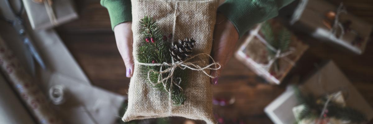 A person's arms, clad in a green sweater, offering up a Christmas gift wrapped in sackcloth, twine, and festive pine needles