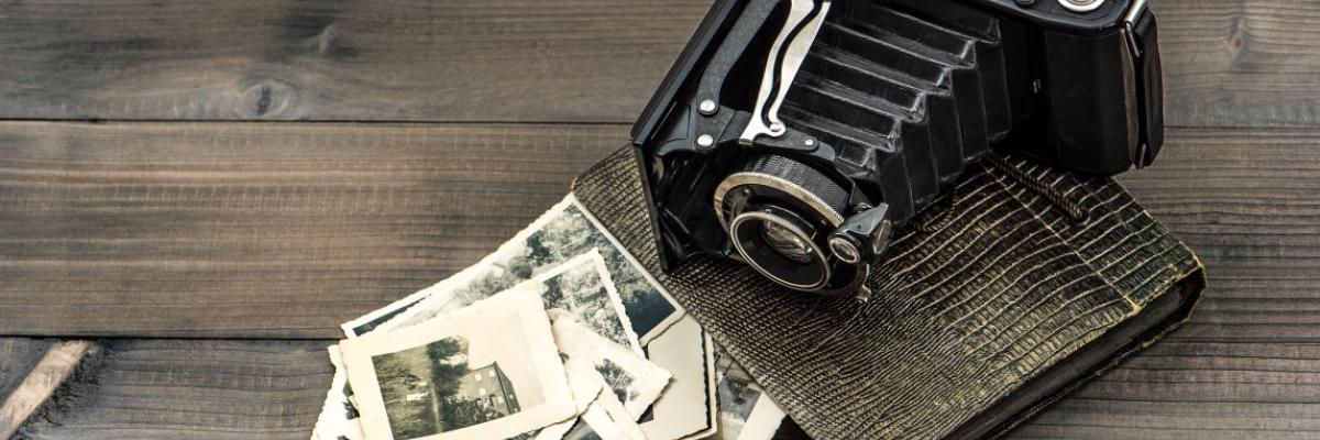 An antique camera lying on a dark brown table, next to a collection of black and white photos