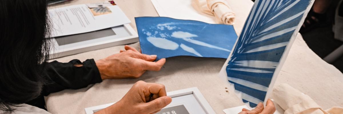 A woman seated at a table, holding up a deep blue cyanotype print in her hand