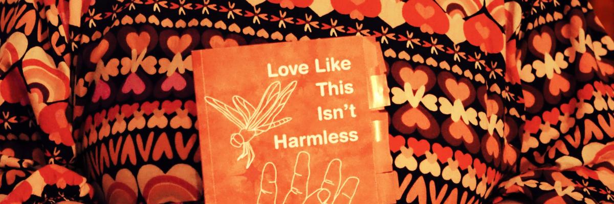A woman wearing a patterned brown, orange, and beige top, holding an orange book titled "Love Like This Isn't Harmless" in her hands