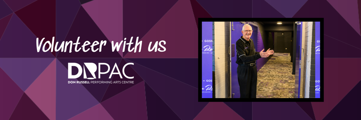 DRPAC volunteer stands smiling next to door way to theatre, gesturing to come in. He is an elderly man with grey hair, wearing glasses, dressed in a black DRPAC-branded shirt with purple piping and logo,  and black pants.