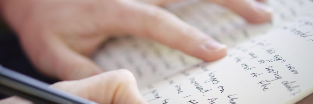 A hand holds a journal open, while the other hand writes words upon the page in pen