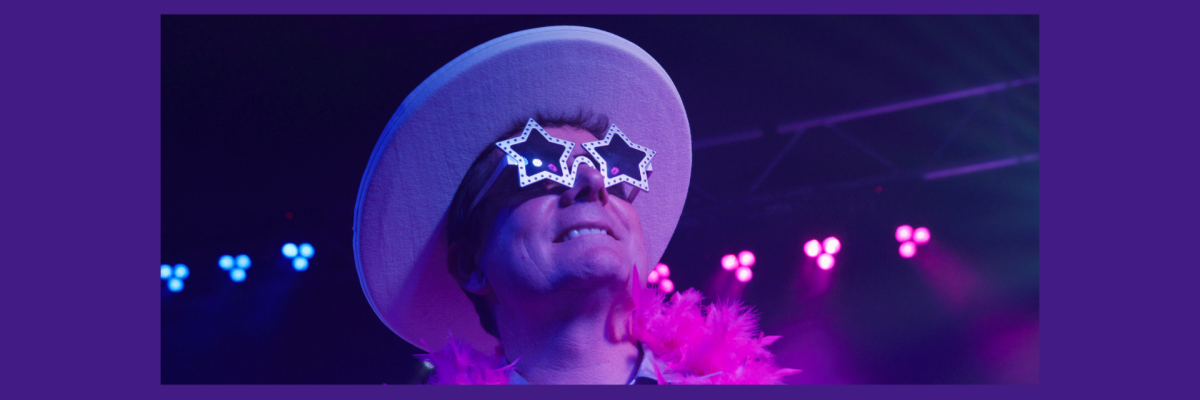 Jay Weston dressed as Elton John stares upwards smiling, wearing a fedora and star shaped white sunglasses.