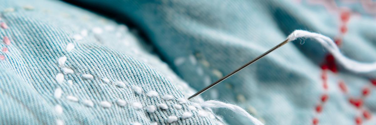 An embroidery/sewing needle, with white thread in its eye, sticking out of denim embroidered with a pattern