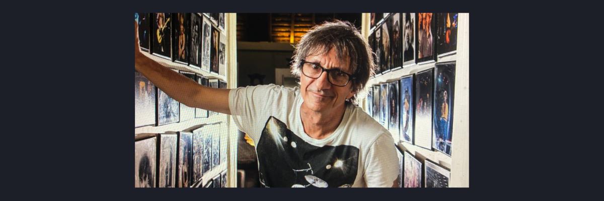 Tony Mott stands in front of a hallway of his photographs, looking at the camera. He wears glasses and a band tee.