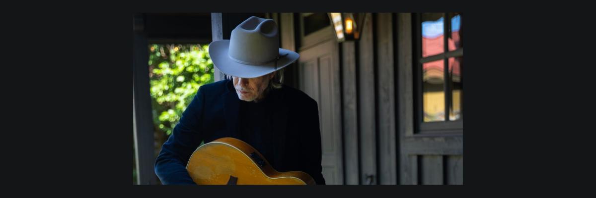 Rick Price stands head down, wearing a fedora and holding an acoustic guitar.