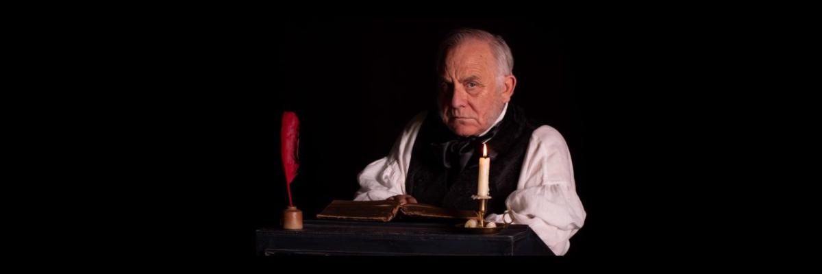 Scrooge sits at a writing desk with a quill, book and candle on it, and stares at the viewer.