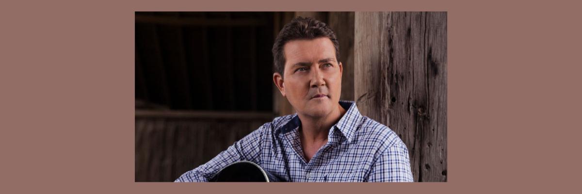 Adam Harvey sits holding an acoustic guitar, looking pensively into the distance.