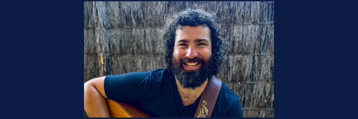 Jeff sits smiling at the camera, with an acoustic guitar on his lap