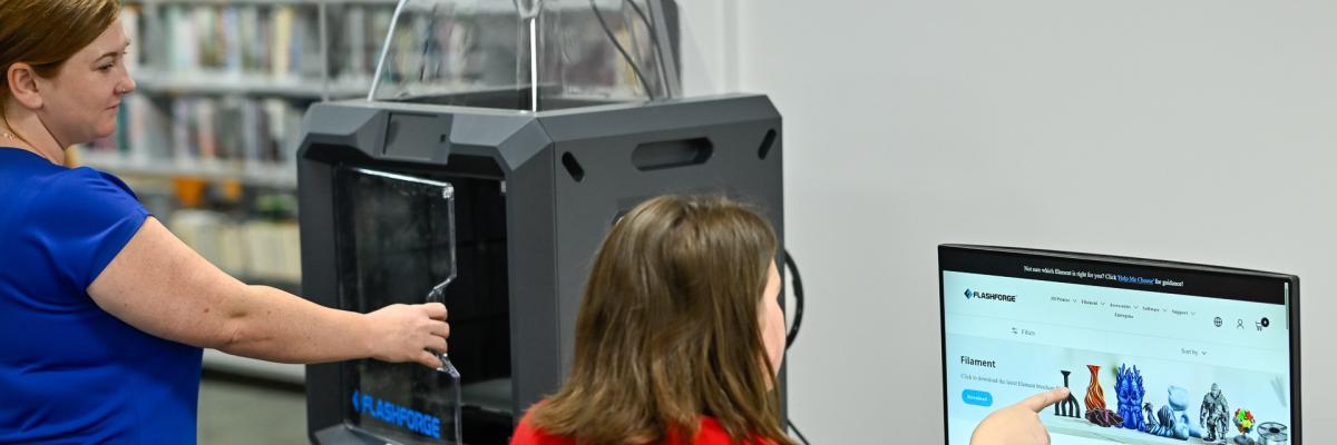 A woman in blue opens the 3D printer door, while a woman in red points at printing designs on the computer screen