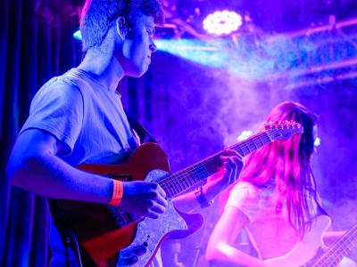 Male Guitarist Playing a red electric guitar surrounded by purple and blue lights shining through haze