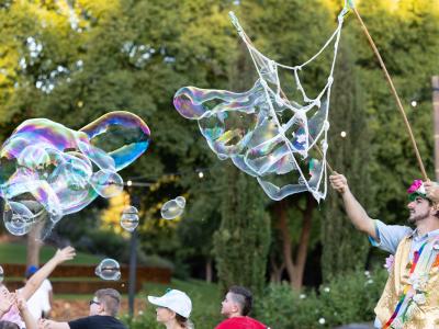 Man dressed up colourfully on the right, blowing big bubbles for kids and adults below to reach