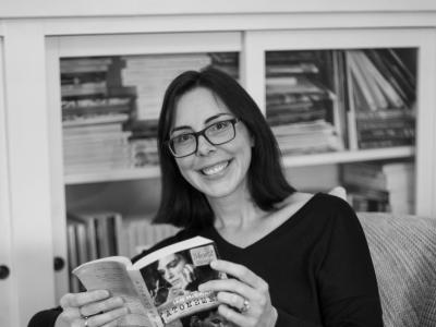 A black and white photo of a woman with a dark bob and glasses, sitting in a chair in front of a book shelf and reading a book.