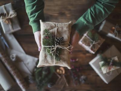 A person's arms, clad in a green sweater, offering up a Christmas gift wrapped in sackcloth, twine, and festive pine needles