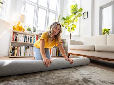 A blonde woman in a yellow shirt and light blue pants, rolling out a carpet in a sunlit room