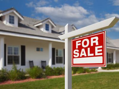 A red and white FOR SALE sign, standing in front of a white house and green lawn