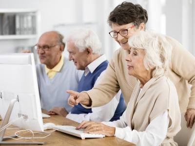 Senior men and women seated in front of computers, hands on keyboards and being taught how to use the technology.