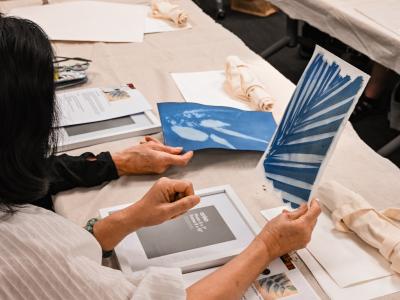 A woman seated at a table, holding up a deep blue cyanotype print in her hand
