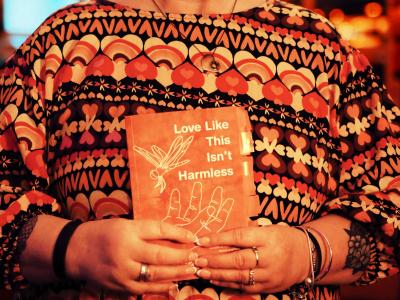 A woman wearing a patterned brown, orange, and beige top, holding an orange book titled "Love Like This Isn't Harmless" in her hands