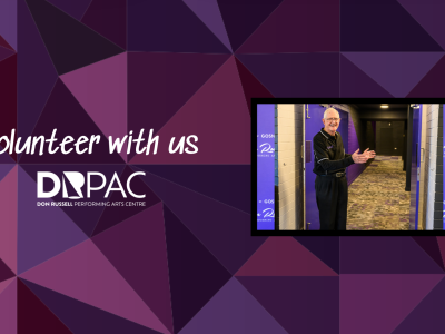 DRPAC volunteer stands smiling next to door way to theatre, gesturing to come in. He is an elderly man with grey hair, wearing glasses, dressed in a black DRPAC-branded shirt with purple piping and logo,  and black pants.