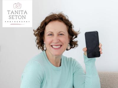 A smiling woman in a pale blue cardigan, holding up a smartphone