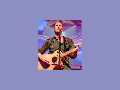 Fred Smith, who is a middle aged white man, stands on stage in front of a microphone, singing and playing acoustic guitar.