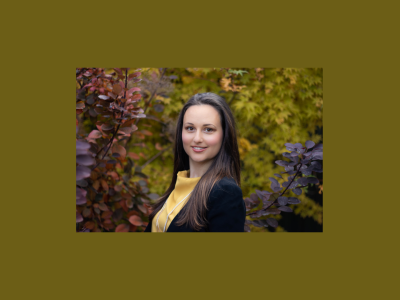 Sarah Waddington stands in front of a backdrop of leaves. Sarah smiles at the camera. She is wearing a yellow shirt.