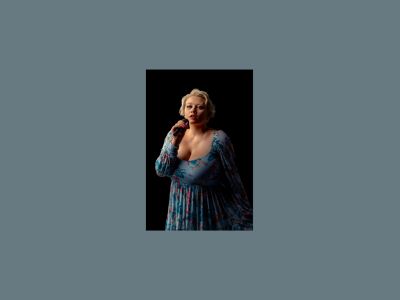 Aria Scarlett stands holding a microphone up to their mouth, looking into the camera. She is wearing a blue floral dress.