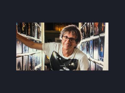 Tony Mott stands in front of a hallway of his photographs, looking at the camera. He wears glasses and a band tee.