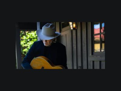 Rick Price stands head down, wearing a fedora and holding an acoustic guitar.