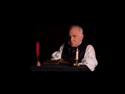 Scrooge sits at a writing desk with a quill, book and candle on it, and stares at the viewer.