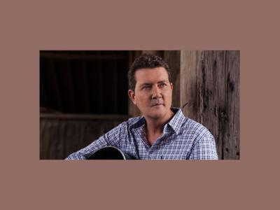 Adam Harvey sits holding an acoustic guitar, looking pensively into the distance.