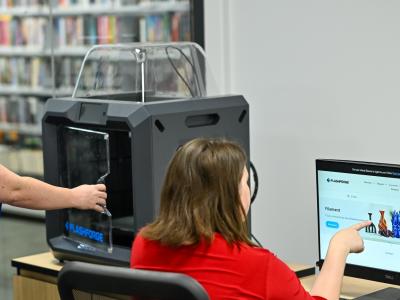 A woman in blue opens the 3D printer door, while a woman in red points at printing designs on the computer screen