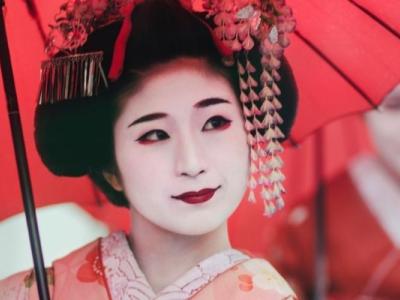 Two geisha in traditional white makeup, dressed in red and pink kimonos, under red parasols